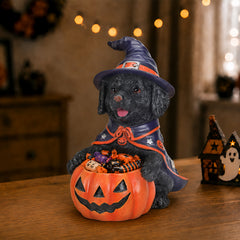Black labrador puppy in a witch hat holding a carved pumpkin candy bowl of treats on a table with candles