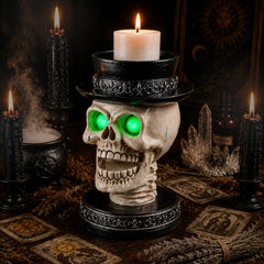 Gothic skull candle holder with a lighted pillar candle in the flat top hat, on a dark table beside cards and a pumpkin