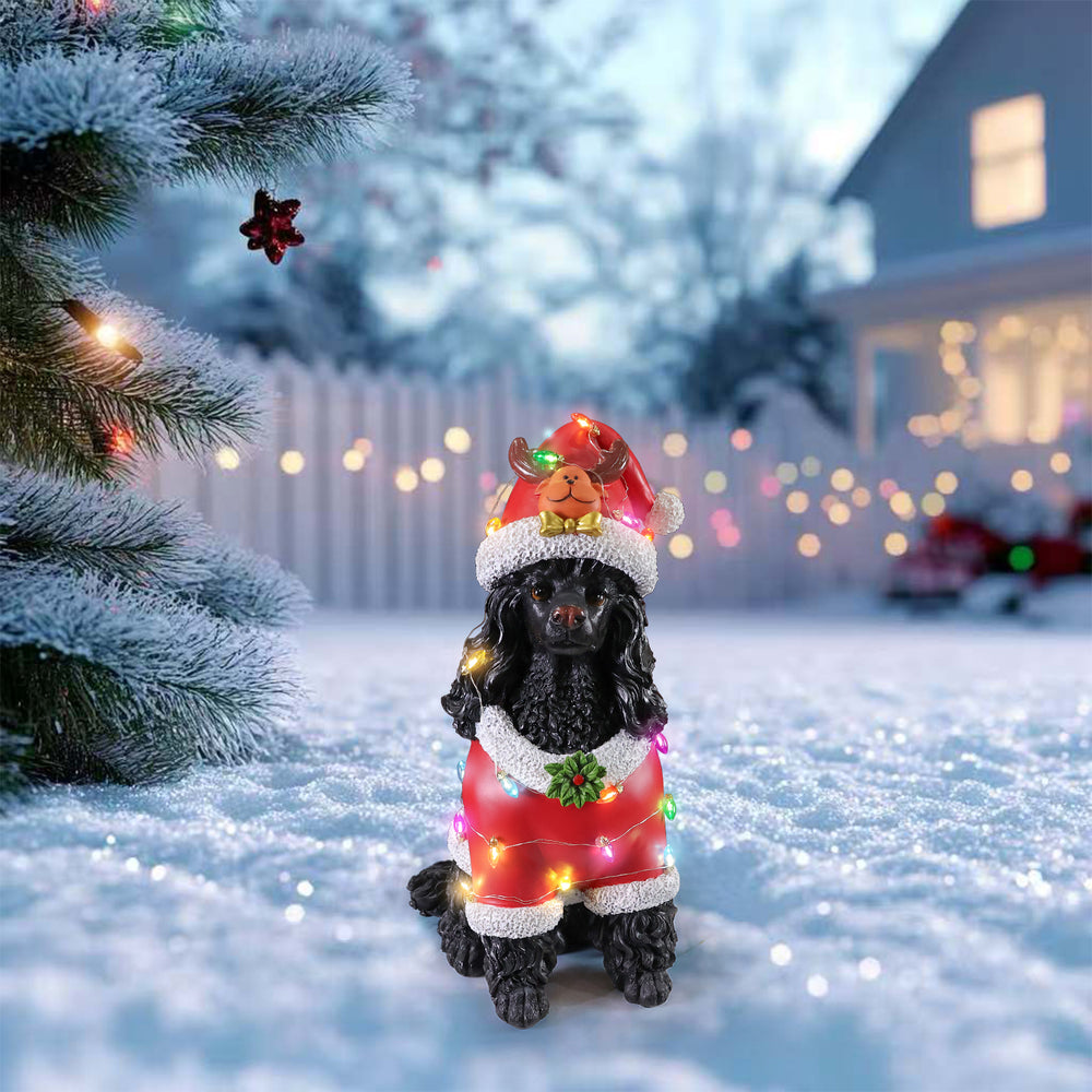 The Black poodle in the red Santa coat standing on snow with a fence and house lights behind