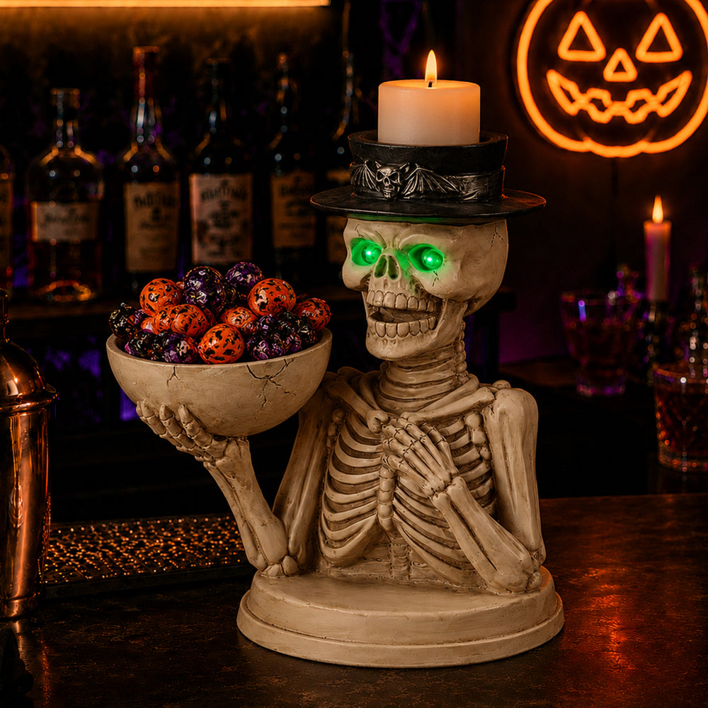 Seated skeleton holding a wide bowl of candy with a black top hat carrying a lit pillar candle on a bar at night
