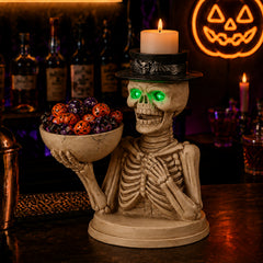 Seated skeleton holding a wide bowl of candy with a black top hat carrying a lit pillar candle on a bar at night