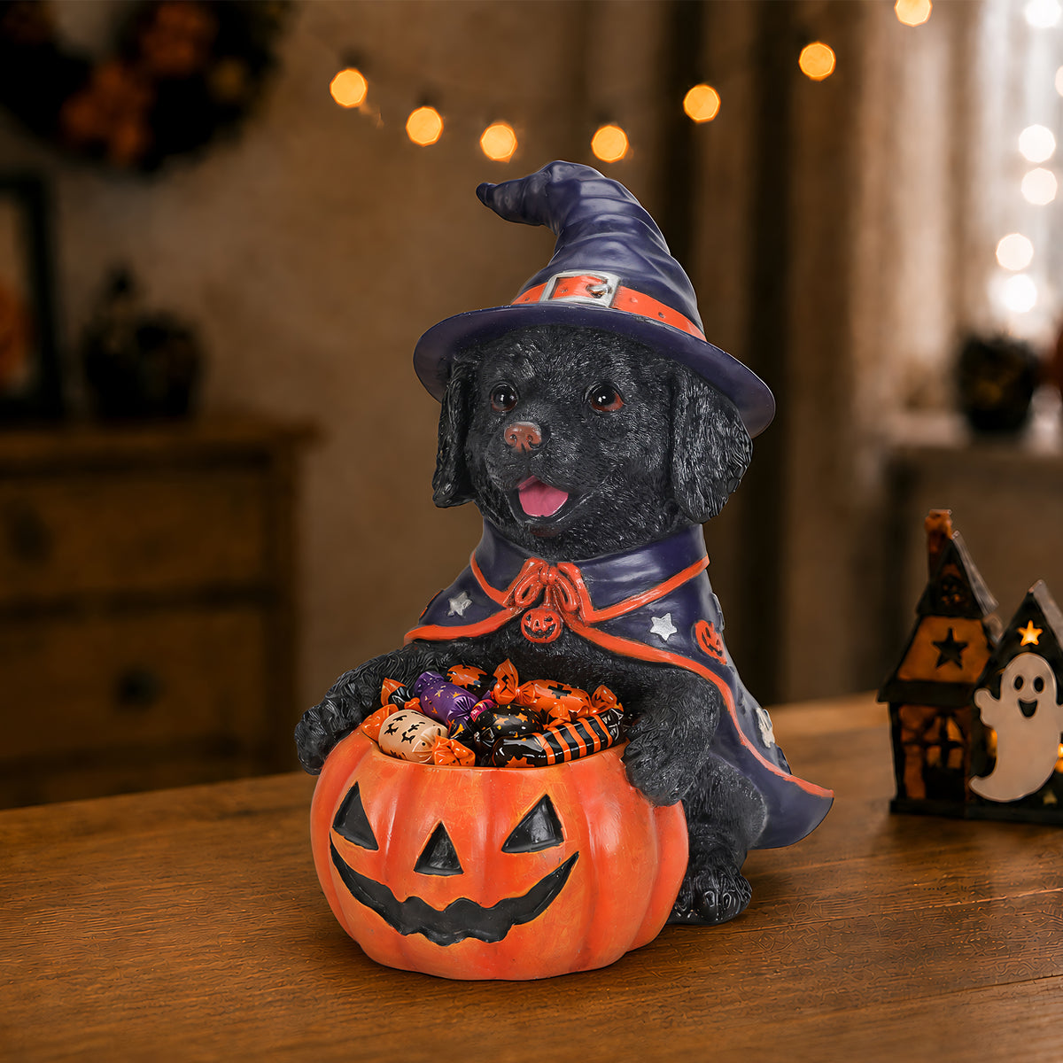 Black labrador puppy in a witch hat holding a carved pumpkin candy bowl of treats on a table with candles