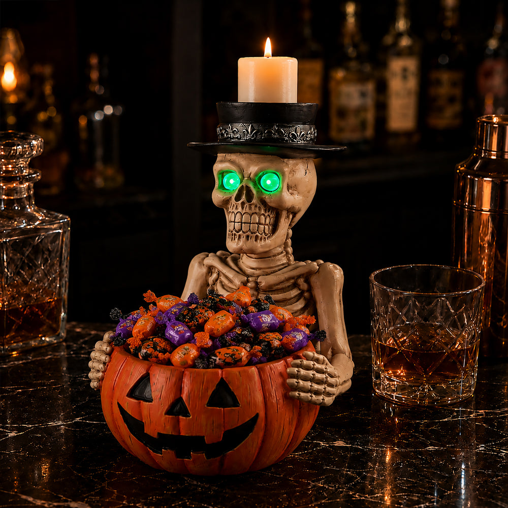 Skeleton figurine holding an orange pumpkin candy bowl filled with chocolates, with a lighted candle standing in its top hat