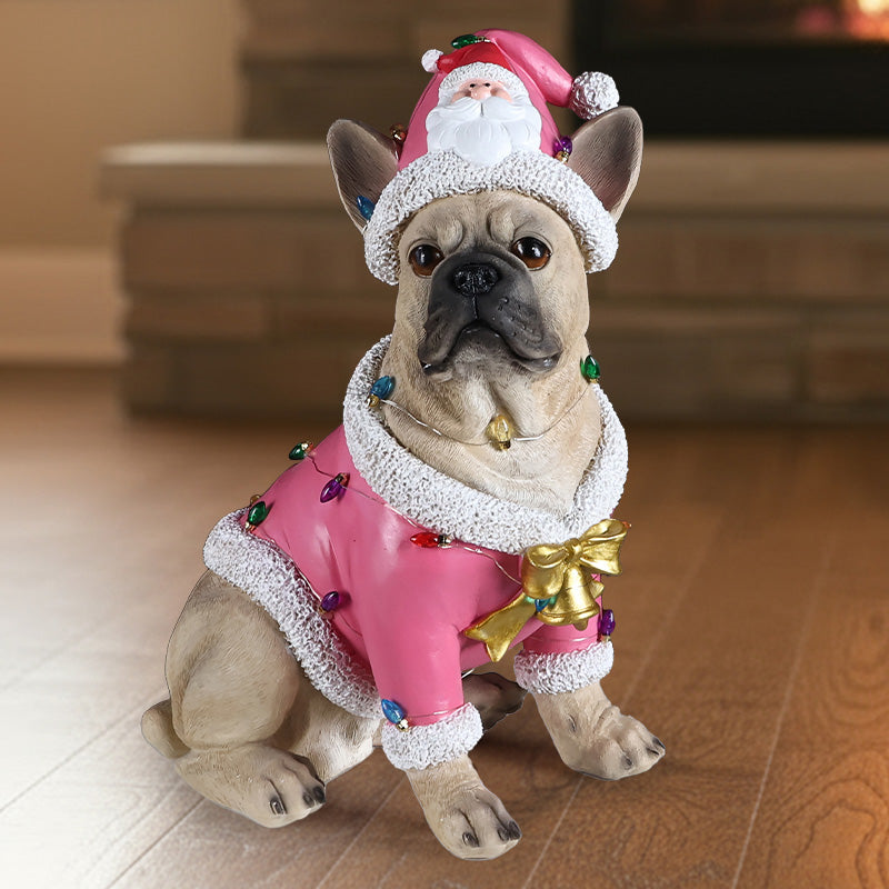 French bulldog in a pink Santa coat with a gold bow and a Santa hat, sitting on a floor in front of a stone hearth