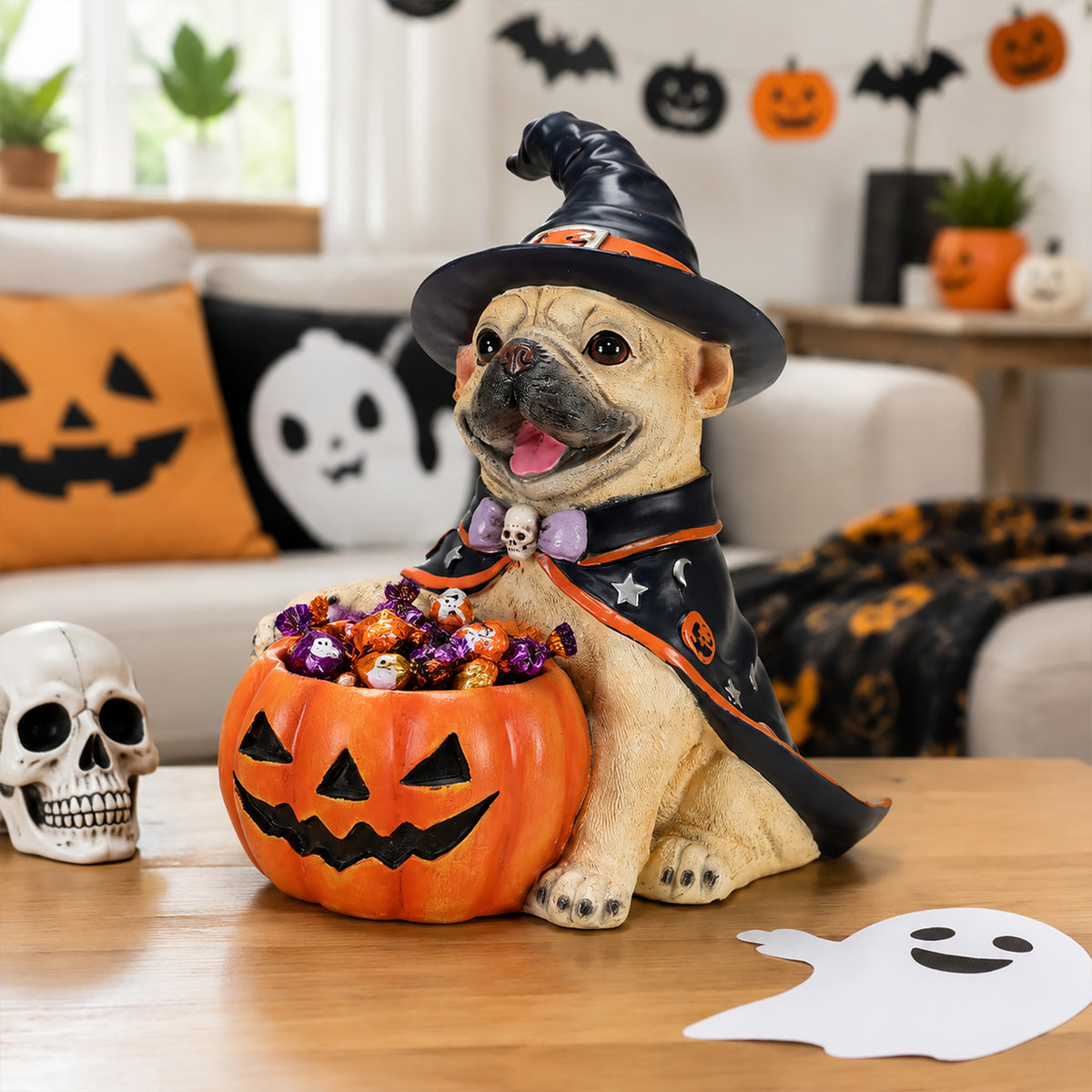 French bulldog witch puppy figure holding a pumpkin candy bowl, on a table with a Happy Halloween sign and a skull beside it